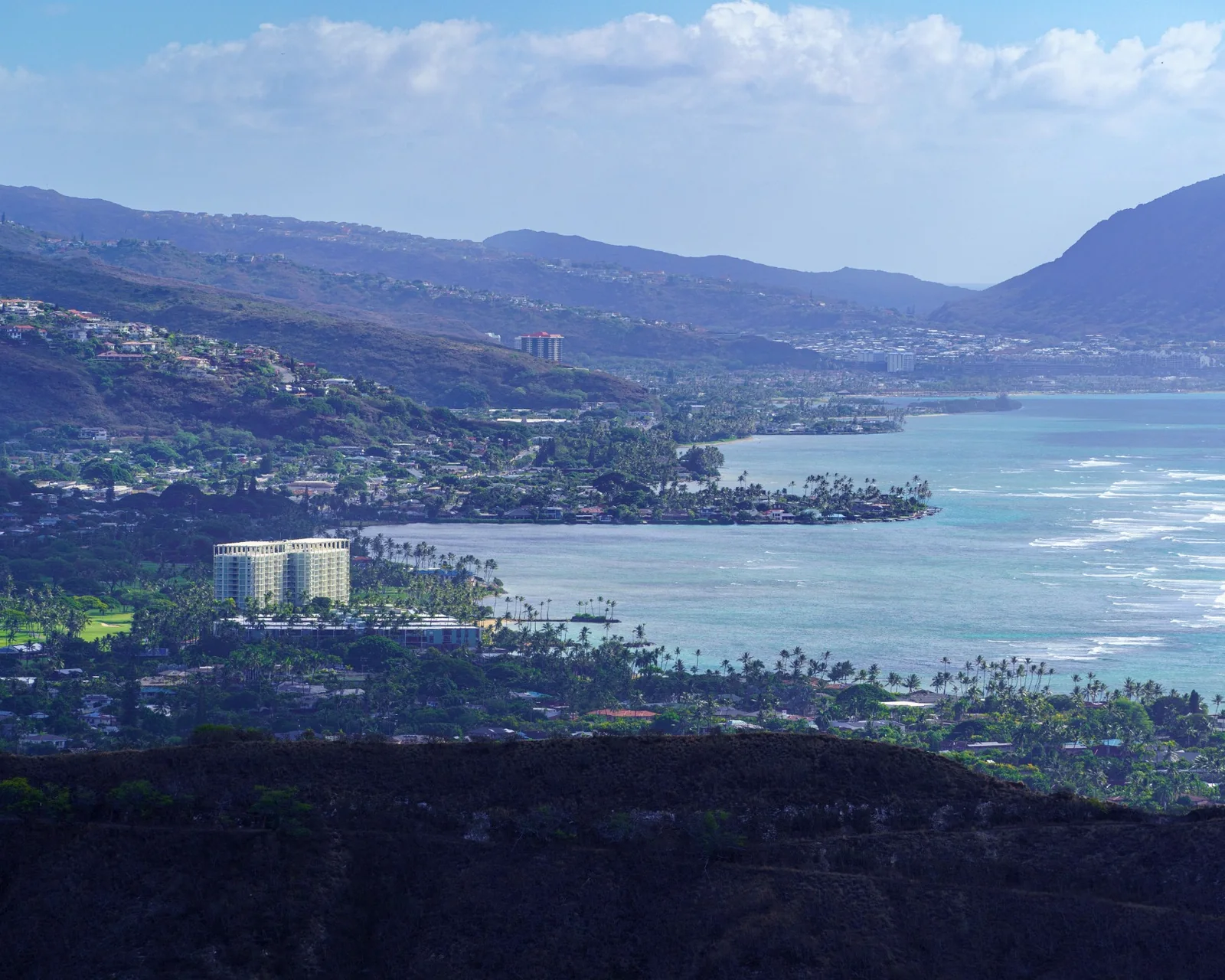 Wide aerial view of Oʻahu neighborhoods, mountain ridges, and a blue coastline beneath scattered clouds