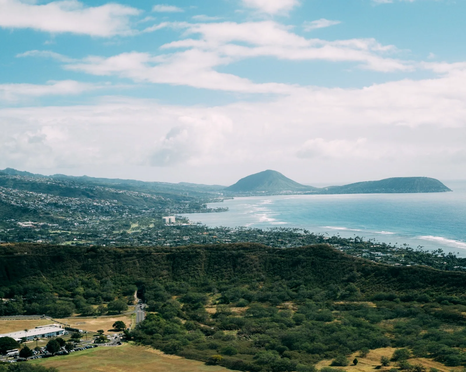 Wide aerial view of an Oʻahu coastline, green ridges, and residential areas under a bright sky